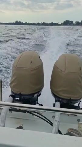 The video shows the wake of a boat moving through water. The view is from the stern of the boat, looking backward. Two outboard motors, covered with tan protective covers, are visible in the foreground. The water is choppy with white foam from the boat's passage. In the distance, a hazy shoreline with trees and some structures can be seen under a cloudy sky. The camera pans slightly to the right, revealing more of the water and the distant land.
