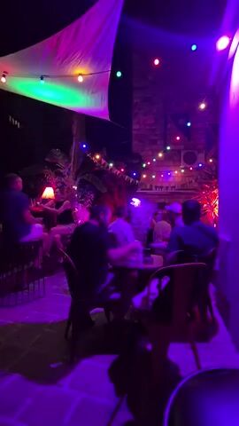 A group of people are gathered outdoors at night. They are seated at tables and a bar, with some standing. The area is decorated with string lights and a large, draped fabric canopy. Purple lighting illuminates the scene. Some individuals appear to be talking and socializing.
