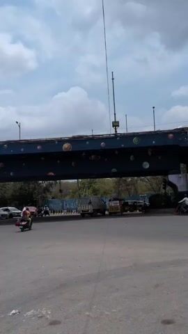 A short video clip shows a street intersection with an elevated highway or bridge overhead. The bridge is painted dark blue with colorful circles. Vehicles, including cars, trucks, and motorcycles, are visible on the road below. A few people are also present, some walking and others on motorcycles. The sky is cloudy.