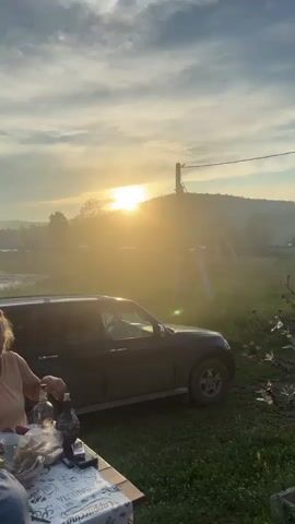 This video captures a group of people gathered around a table outdoors during sunset. The sun is low in the sky, casting a warm, golden light and creating a hazy atmosphere. In the foreground, a black SUV is parked. A woman with blonde hair is seated at the table, appearing to prepare food. There are various items on the table, including bottles, bread, and vegetables. Further in the background, a body of water is visible, with a distant shoreline featuring houses and trees. Another man wearing a hat is also seated at the table. A third man, wearing a white t-shirt, stands near the table. The overall scene suggests a relaxed, informal gathering in a rural or semi-rural setting.