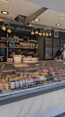 The video shows the interior of a store filled with cheese and other food items. In the foreground, a display case is filled with various types of cheese, some of which are labeled. Behind the display case, there are shelves stocked with jars, bottles, and packaged goods. A meat slicer is visible on a counter. The store appears to be a deli or specialty food shop.