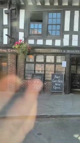 The video pans across a street with a row of buildings. The first building appears to be a pub or restaurant with outdoor seating and chalkboards advertising lunch specials and live music. The second building is a shop with a white facade and large windows displaying merchandise. The third building is a charity shop with clothing displayed in the windows. Several people are visible on the sidewalk, including one person kneeling on the ground. The overall environment appears to be a commercial street in a town or city.