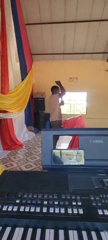 The video appears to be taken inside a simple, indoor space that could be a small church or community hall. A man is standing behind a table, speaking into a microphone. He is wearing a light-colored shirt and brown pants. Behind him, there's a window and some draped fabric in red, yellow, white, and blue. To the left, there are speakers and a piece of furniture. In the foreground is a Yamaha keyboard, with a smartphone placed on top of the keyboard's display. In the background, to the right of the speaker, there are several people seated in blue plastic chairs, seemingly listening to the man speak. The room has a patterned tile floor, pale yellow walls, and a metal truss roof.