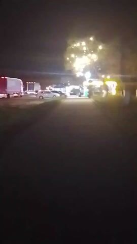 The video shows a dark outdoor scene, seemingly at night. The foreground is a dark path. In the distance, there is a cluster of vehicles, including what appears to be a semi-truck. There are also some structures lit by bright lights. Some of the lights are strung up on a tree. There are a few people visible in the distance. The overall atmosphere is dark, with only the lights from the vehicles and structures illuminating the scene. The footage is slightly shaky.