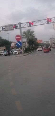 The video presents an active urban street intersection in Hong Gai Ward, Vietnam, under an overcast sky. The scene is dominated by a mix of vehicles, including cars (sedans, SUVs, and a truck/van) and numerous motorbikes, which are either stationary or in motion.

Overhead traffic lights are prominently displayed, showing red signals with digital countdown timers indicating remaining seconds, such as '89', '84', and '82'. A central street pole at the intersection features two circular traffic signs: a red "No Entry" sign (red circle with a white horizontal bar) and a blue "Mandatory Turn Right" sign (blue circle with a white arrow pointing right and slightly downwards).

The camera briefly pans to capture the movement of a black SUV driving through the intersection. Multi-story buildings line the streets, some appearing to be commercial establishments, and large billboards with Vietnamese text are visible on their facades. One billboard clearly shows "BAO VIET" and "SO THAI" among other texts. Palm trees and other sparse greenery adorn a central median and sections of the roadside. The road is paved with asphalt, and faint pedestrian crosswalk markings are present. The overall activity depicts typical urban traffic flow, with vehicles waiting at red lights and proceeding when clear.