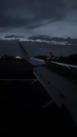The video shows a view from an airplane window at night. The wing of the plane, which has "RYANAIR" printed on it, is visible. The plane is on the ground, possibly on a runway.  There are lights and some buildings in the distance. The sky is dark and cloudy. The overall atmosphere appears dim and somewhat moody.