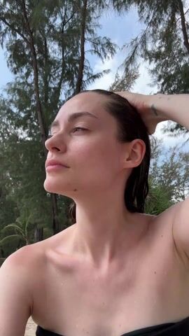 The video shows a woman outdoors, with a partial view of her bare upper body. She initially stands with her eyes closed, head tilted slightly upward. The background features tall trees and a sky with scattered clouds. She then raises her right arm and places a green baseball cap on her head, adjusting it slightly. Her right arm bears tattoos of an umbrella and a constellation. The overall impression is one of a casual, possibly beach-related scene.