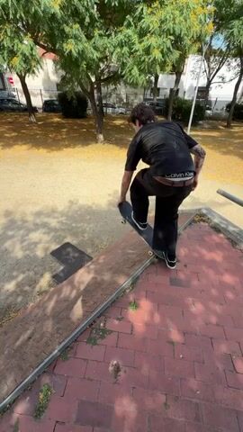 A young man with tattoos on his arm wearing a black t-shirt and black pants is skateboarding on a brick ramp. He is in the process of performing a trick as he is in mid-air. The skateboard is in motion. The man is outside in a park-like setting with trees and gravel on the ground. The sun is shining and casting shadows. He is looking to his right. The video appears to be shot in a vertical orientation.