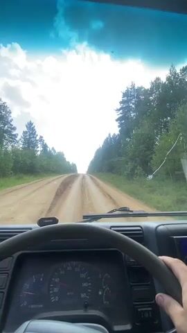 This video shows the interior of a truck cab as it drives on a dirt road. The view is from the driver's perspective, looking out the windshield and side window. The dashboard is visible, showing a speedometer and tachometer. The road is surrounded by trees. The sky is cloudy with bright patches of sunlight. The truck is moving forward on the dirt road.
