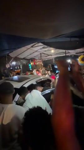 A crowd of people are gathered around a white car at night. Many people are holding up their cell phones, some with flashlights on, to take pictures or videos. Some individuals are reaching towards the car, and one person is leaning into the open driver's side door. The environment appears to be an outdoor market or street area, with some structures and tents visible in the background, illuminated by artificial lighting. There is a sense of excitement and activity.