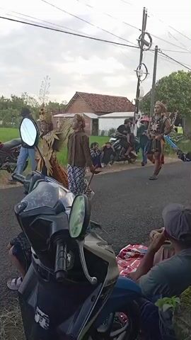 The video captures a scene of people gathered on a narrow, paved road in a rural or suburban setting. Several children are dressed in costumes that appear to be part of a cultural performance or parade, with ornate headpieces and traditional clothing. One of the costumed performers is walking down the road, while others are sitting or standing around. In the foreground, a parked black and white motorcycle is visible, with a man sitting nearby, resting on a blanket. Across the road, there's a view of a rice field and some houses in the background. Utility poles with wires stretch across the sky above the road. The overall atmosphere seems relaxed and communal, indicative of a local event or gathering.
