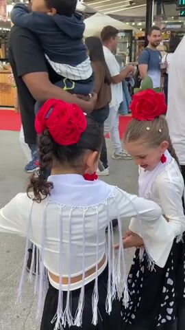 Two young girls in matching flamenco dresses and red flower headpieces are dancing in a crowded outdoor market. The girl on the left has a long braid while the other has a long braid with flowers weaved in. They smile as they perform a simple choreography, spinning and moving their arms. They are surrounded by people browsing stalls with food and other merchandise. The ground is paved, and there is a red carpet nearby. The atmosphere is festive and lively.