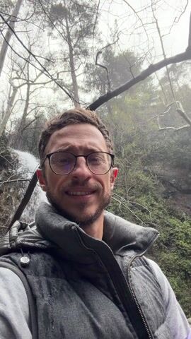 A man wearing glasses and a gray vest smiles at the camera. He is outdoors in a forest-like area with trees and a waterfall behind him. The weather appears to be damp, and some bare branches are visible. He holds up a shaka sign with his right hand.