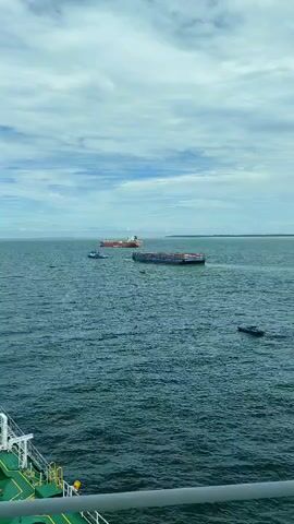 A video shows a wide shot of a body of water, likely the sea, under a cloudy sky. In the distance, several ships are visible. One large orange and white tanker is sailing away from the camera. In front of it, a long barge filled with shipping containers is being pushed by a smaller blue tugboat. To the right of these vessels, a small dark boat is moving across the water. In the foreground, the railing and deck of a ship are visible, indicating the camera is positioned on board a vessel. The water is a deep blue-green and appears somewhat choppy.