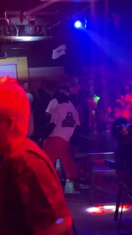 The video captures a lively scene inside what appears to be a nightclub or bar, filled with people socializing and dancing. The lighting is dim, dominated by pulsating red and blue stage lights, with vibrant green laser beams sweeping across the room, creating an energetic atmosphere.

In the foreground, a person with bright red hair is partially visible, facing towards the camera. The main focus is on a group of people gathered in the center and background. One person in particular stands out, dancing actively in the middle, wearing a white t-shirt with a gold logo (featuring a crown and the letter 'B', possibly "BOSS BAR") and bright orange pants. Another individual is seen wearing a green beanie with text, possibly "JESU".

Tables and chairs are scattered throughout the venue, some with bottles on them, indicating a social gathering. The crowd appears to be enjoying the music, which is audible in the background, contributing to the festive and upbeat mood of the event.