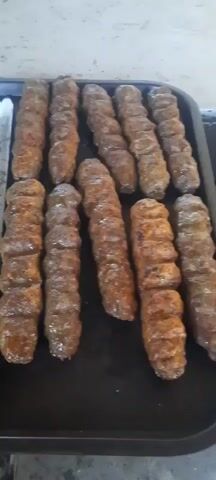 This video shows rows of freshly grilled meat skewers on a metal tray. The skewers are arranged in parallel lines, with some appearing to be more cooked than others. Metal tongs are visible on the left side of the frame, suggesting that the skewers are being prepared for serving or are in the process of being cooked. The meat appears to be finely ground and has a seared, browned exterior, indicating it has been cooked over heat. The tray is placed on a surface that seems to be a grill or a cooking station. The lighting is natural, and the focus is on the food.