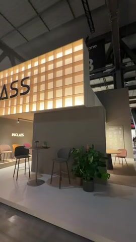 The video provides a sweeping view of the "INCLASS" exhibition booth, likely situated within a large convention or trade show hall in Rho, Italy, indicated by the industrial ceiling structure.

The booth's facade is dominated by a large, illuminated sign displaying "INCLASS" in prominent black letters against a backdrop of glowing, grid-patterned panels. Below this sign, the entrance area features a small, informal seating arrangement with two grey upholstered bar stools and a circular high-top table. A dark potted plant adds a natural accent next to one of the stools.

Stepping into the booth, a spacious interior area is revealed on a raised, light-colored platform. This section is designed as a contemporary living space, showcasing a variety of "INCLASS" furniture. Comfortable sofas and ottoman-style seating in neutral tones of beige and grey are arranged to create inviting lounge areas. Along one wall, a display highlights individual chairs in a vibrant palette of colors including muted pink, bright orange, sunny yellow, and deep green.

Several people are visible inside the booth, moving casually, observing the furniture, and interacting with each other, suggesting a typical trade show atmosphere where attendees browse and learn about products. The booth is brightly lit with overhead spotlights and ambient backlighting from the main sign, creating a welcoming and modern ambiance. The interior walls are a consistent, neutral grey-brown, allowing the diverse furniture designs and colors to stand out prominently.