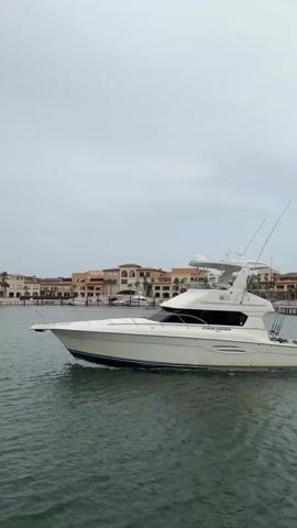 A white yacht with a dark blue stripe along its hull is cruising on calm water. The yacht has a raised cabin with dark tinted windows and a flybridge. Two fishing rods are mounted on the stern. A person wearing a blue shirt and white shorts is visible on the deck near the stern. In the background, a row of Mediterranean-style buildings with terracotta roofs lines the shore. The sky is overcast.