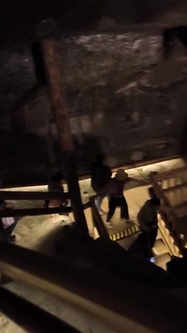 The video depicts a dimly lit, possibly underground, structure with rough, rock-like walls. A series of wooden ladders or platforms are visible, ascending upwards out of view. Figures of people can be seen indistinctly climbing or standing on these structures, silhouetted against a lighter background. The camera appears to be positioned at the base, looking upwards.