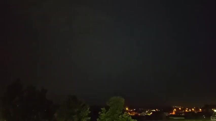 The video captures a nighttime view of a residential area under a dark sky. In the foreground, silhouetted trees partially obscure the view of houses with illuminated windows and streetlights. The sky is mostly dark, but a bright flash of lightning illuminates the clouds and the landscape for a brief moment. This flash appears to be a cloud-to-ground or intra-cloud lightning strike, casting a strong white light across the scene. Following the flash, the sky returns to its dark state.