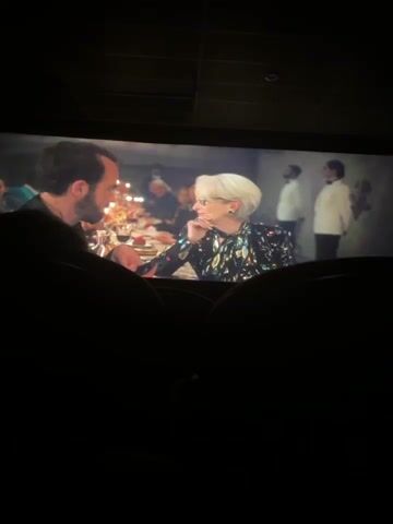 The video captures a scene from a movie being watched in a dark movie theater. The audience is mostly obscured in the foreground, with only silhouettes of their heads and shoulders visible. On the movie screen, a dinner scene unfolds. In the initial frame, a woman with shoulder-length auburn hair, wearing a black dress with a silver necklace, is seated at a table, smiling. As the scene shifts, it focuses on an older woman with short white hair and glasses. She wears a sparkly black jacket, and is also seated at the table, smiling.  A man with a beard sits across from both women, partially visible on the left side of the screen. The background reveals other individuals at the dinner table, along with flickering candlelight, creating a warm ambiance.