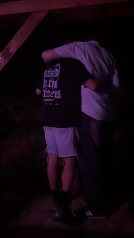 Two individuals stand in a dark environment, illuminated by purple light. The person on the left wears a black t-shirt with white text on the back and light blue frayed denim shorts, with their back partially to the camera. The taller person on the right wears a light-colored short-sleeved collared shirt and dark pants, with their back and right side towards the camera. The taller individual has their right arm wrapped around the shoulder of the person on the left, who appears to be embracing them back. Both individuals' faces are obscured. Background music is audible.
