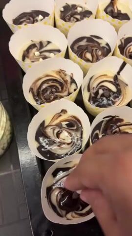 The video shows a close-up, top-down view of a person's hand adding white chocolate chips to several unbaked cupcakes. The cupcakes are arranged in white paper liners with yellow polka dots, placed on a dark surface. The cupcake batter has a swirled pattern of white and dark chocolate. The person's hand is in the foreground, and they are dropping small white chips into the batter of the cupcakes. There is a jar of white chocolate chips to the left of the cupcakes. The lighting is bright, and the focus is sharp.