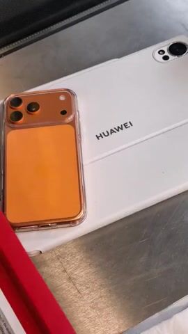 The video shows a close-up shot of a white Huawei tablet and an orange-colored phone case, possibly for an iPhone. The phone case is placed on top of the tablet, obscuring a portion of the Huawei logo. The tablet is lying flat on a metallic surface, which appears to be a countertop. The lighting is bright, and the focus is on the objects. There are no people visible in the video. The scene is static, with no apparent action or interaction occurring.