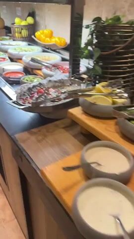 A buffet salad bar is shown. It features various dishes including salads with leafy greens, chopped vegetables like tomatoes, cucumbers, and carrots, as well as pasta dishes, beans, and dips. There are also whole fruits like lemons and bell peppers displayed in baskets and bowls. The food is presented in white and grey bowls on a dark countertop, with tongs provided for serving. The background shows a tiled wall and wooden cabinetry. The camera pans across the buffet, offering a close-up view of the food options. No people are visible in the video.