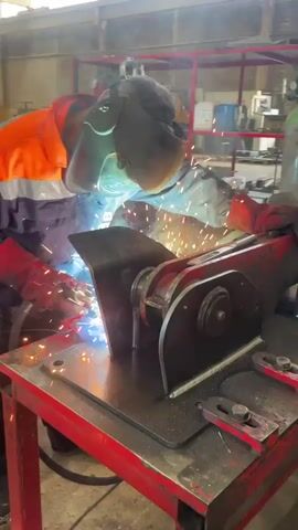 In an industrial setting, a welder is actively welding a metal object placed on a red metal table. The welder, wearing safety gear including a welding mask, red gloves, and a hi-vis orange jacket over a long-sleeved shirt, is focused on the welding process. Sparks are emitted from the point of contact between the welding torch and the metal, illuminating the immediate area. The metal object being welded is attached to a larger, red mechanical structure. Background elements include gas cylinders and other metal equipment, suggesting a workshop or factory environment.