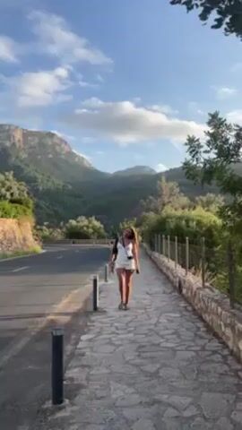 A woman walks along a stone sidewalk next to a road with mountains in the background under a partially cloudy sky. She wears a white top and shorts and carries a black bag. A cyclist rides past her on the road, wearing a red and blue shirt and black shorts. The cyclist speeds past the camera.