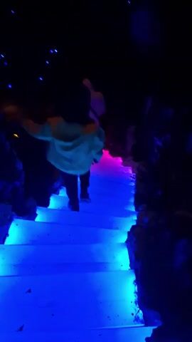 The video shows a person walking down a set of illuminated stairs. The stairs change color from blue to pink to red as they descend. The person is wearing dark pants, black shoes, and a light-colored jacket or coat. There is a second person, further away and appearing as a silhouette, also moving in the same direction. The stairs are lined with rocks or cave-like formations on either side. The lighting is dark, enhancing the color contrast of the stairs. The environment is enclosed, resembling a cave or tunnel.