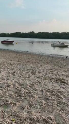 The video depicts a scenic view of a riverbank at sunset. The foreground is dominated by a sandy beach, with scattered pebbles and some sparse vegetation. The water is calm and reflects the warm hues of the setting sun. Several boats are visible on the river, including a white pontoon boat in the middle ground and a red and white personal watercraft further to the left. In the distance, a dense line of trees forms the horizon, and a tall electricity pylon stands out against the sky. The sun is setting behind the trees, casting a golden glow over the scene and creating a warm, tranquil atmosphere. The sky is a mixture of pale blue and orange, with a few wispy clouds. There are no people visible in the footage. The overall impression is one of peaceful natural beauty, likely captured during a summer evening in Dunakeszi, Hungary.