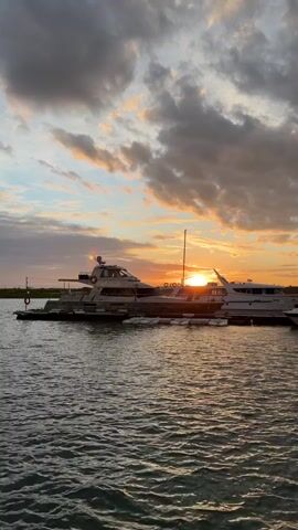 The video shows a serene harbor at sunset. Several yachts are docked, their white hulls reflecting the warm glow of the setting sun. The sky is a canvas of dramatic clouds, tinged with orange, pink, and yellow hues from the sun peeking through. The water is calm, with gentle ripples catching the light. The scene evokes a sense of peace and tranquility.