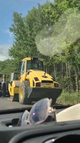 The video shows a construction scene on a road. It begins with a close-up of a yellow steamroller. Then, the view shifts to a group of construction workers standing around a large piece of equipment, possibly a dump truck or a trailer. One worker is wearing a high-visibility vest. The next shot shows the back of a white van with its doors open, suggesting it is part of the construction operation. The video then pans to the road ahead, showing a white van and an orange steamroller on the road. The road appears to be under construction, with areas of fresh asphalt. The surrounding environment is rural, with trees and hills visible in the background under a partly cloudy sky. Sunglasses are visible in the foreground of the first three shots, indicating the video is likely filmed from inside a vehicle.