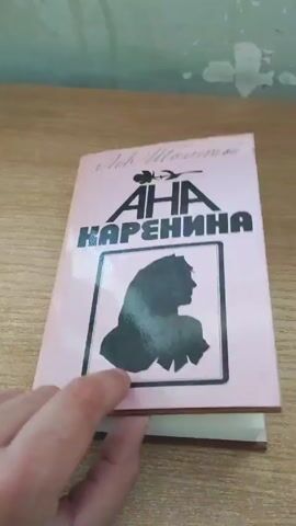 A hand is turning the pages of a book titled "Anna Karenina" by Leo Tolstoy, volume one. The book is in Bulgarian, translated by Ivan Jechev, and published by "Raduga" in Moscow and "Narodna Kultura" in Sofia.  The video is filmed on a wooden surface.  The book is hardcover. The pages are white.
