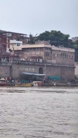 The video shows a view of a building complex by a body of water. The buildings appear to be made of stone and have multiple levels and architectural features, including what looks like a temple with red spires. There are stairs leading down to the water. Several boats are visible on the water, and some people are seen near the stairs and on the boats. There are also some cloths hanging on a line. The water appears to be murky and brown. The sky is overcast. There is a sign on the building that reads "BANARAS GHAT."