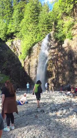 The video captures a scenic view of a waterfall cascading down a rocky cliff face, surrounded by lush green trees. A group of people are present near the base of the waterfall and along the rocky riverbed. Some are sitting on the ground, while others are walking or standing. One person is seen walking towards the waterfall, wearing a green backpack. Another person is seated on a rock, looking at their phone. The river flows over a bed of smooth stones. A bridge is visible overhead in some shots, spanning the gorge. The overall atmosphere appears relaxed, with people enjoying the natural setting.