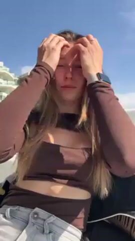 A young woman sitting outside on a sunny day. She is wearing a long-sleeved brown shirt with cutouts in the front and light-wash jeans. She has long, light brown hair. She is looking up and touching her face, and then briefly looks off to the side. There are buildings in the background and a bright blue sky.