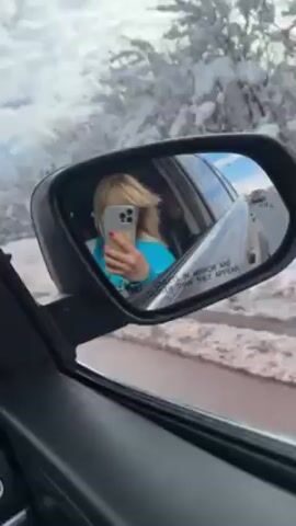 A car is driving along a snowy road lined with snow-covered trees. The view is primarily through the driver's side mirror, showing a woman with blonde hair wearing a blue shirt taking a selfie with her smartphone while driving. The mirror's reflection also shows the car's interior, part of the steering wheel, and other vehicles on the road. The road is partially covered in slush and snow. The sky is overcast. The mirror has a safety warning printed on it: "Objects in mirror are closer than they appear." The footage appears to be taken from inside the car.