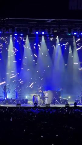 The video shows a live concert with a stage lit by bright blue spotlights. Confetti is falling from above. On stage, several musicians are performing. In the foreground, an audience is visible, with many people holding up their phones. A large screen behind the performers displays abstract patterns and a faint image of a person's face. One performer, a bald man in a white t-shirt and white pants, is at a keyboard and appears to be singing or speaking into a microphone. He moves around the stage and interacts with the equipment.