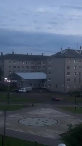 A static, slightly shaky, vertical video captures a view of an apartment complex and its surrounding area at dusk. The sky is overcast with visible clouds. In the foreground, a circular paved area with a decorative pattern of dark circles on a lighter background is visible. A dark pole stands to the left of this area. Beyond the paved area is a street with a white van parked to the left of a low-rise building attached to a larger apartment building. A dark-colored car is driving away from the viewer on the street, its brake lights illuminated. The apartment buildings are multi-story structures with visible windows. Lights are visible in some of the windows, and a brightly lit entrance area is situated between two sections of the buildings. There are trees and grass visible to the left of the apartment complex. No people are clearly discernible in the video.