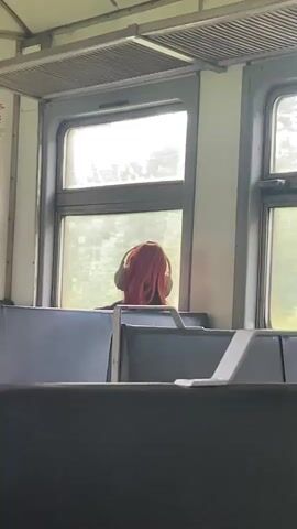 A medium shot shows the back of a person with bright red hair wearing headphones and looking out a window on a train. The person is seated on a gray fabric bench. The train car interior is visible, with other gray benches and a metal luggage rack overhead. The window shows an out-of-focus view of greenery outside. The lighting is bright and appears to be daylight. The camera is stationary.