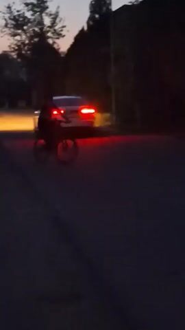 A person on a bicycle rides away from a car with its brake lights illuminated in the dark. The car is positioned to the left of the frame, while the bicyclist is in the center. The bicyclist is moving away from the car and out of the frame to the right. The scene is dimly lit by the car's lights and possibly ambient light from the sky. The surroundings are obscured by darkness, with some indistinct shapes that may be trees or foliage visible on the right side of the frame. The road appears to be asphalt.