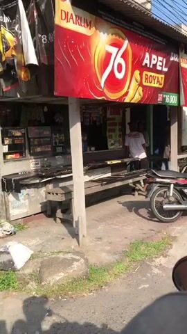 The video shows a street scene outside a small shop. A red and yellow banner hangs above the shop, advertising "APEL ROYAL" and displaying a price of "RP 16.000". In front of the shop, there are several wooden planks laid out, possibly for displaying goods or as seating. A motorcycle is parked in the foreground. Two individuals are visible near the shop, one in a white t-shirt with red writing and another person further back. Various items are displayed on shelves inside the shop. The environment appears to be outdoors, with sunlight indicating daytime.