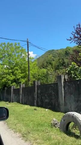 The video presents a scenic view captured from a moving vehicle on a bright, sunny day. The scene unfolds across a lush green landscape, suggesting a rural or semi-rural setting, consistent with Achadara, Abkhazia.

The primary subjects include a variety of green trees and dense vegetation covering hills that rise in the background. A clear, vibrant blue sky with scattered white clouds dominates the upper portion of the frame.

In the foreground, a gray concrete wall topped with a black wrought-iron fence is visible, followed by a distinctive green metal fence featuring a repeating diamond-shaped pattern. Several utility poles, both wooden and concrete, with associated power lines, are interspersed throughout the view. One of the concrete poles also holds a street light. Small, block-like structures are partially visible on the green slopes. The side mirror of the car is visible in the bottom left corner, indicating the perspective from inside a moving vehicle.

The activity shown is the continuous panning of the landscape as the car drives along a road. The time of day is clearly daytime with bright sunlight, and the weather appears clear and pleasant. No discernible text is visible within the video frames.