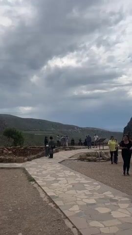 A panning video captures a sunny outdoor scene, starting with a view of a stone pathway where people are strolling. The pathway is lined with low stone walls and leads towards a large, rugged mountain. A building with a conical roof is visible at the base of the mountain, surrounded by stone ruins. A group of people are gathered on the pathway, some walking towards the camera, others standing and observing the surroundings. The sky is partly cloudy, with patches of blue visible between the white and gray clouds.