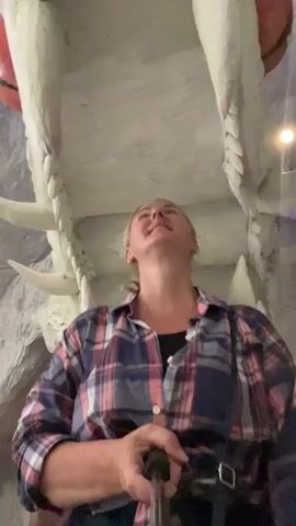 A woman in a plaid shirt is holding a selfie stick and looking up at a large, white, dragon-like sculpture with sharp teeth. The sculpture appears to be a representation of an open mouth. The environment is dimly lit and seems to be an indoor exhibit or attraction. The woman is smiling and seems excited as she records the video.