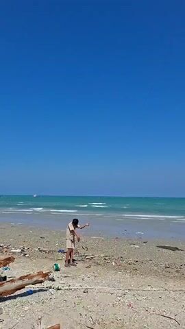 A person stands on a beach, holding a kite string. The kite, white and shaped like a jellyfish, flies high in the bright blue sky. The ocean is visible in the background with small waves. There is some debris on the beach.