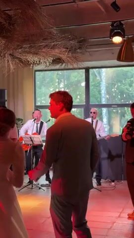 A wedding reception is underway as a wedding band plays live music on a stage. The band consists of three men. One man is singing and playing a red electric guitar. Another man is playing an electric guitar and singing, while a third man is behind a drum set. The band members are dressed in white shirts and black pants. The guests are dancing and enjoying the music. A man in a suit is facing the band, his back to the camera. A woman in a white wedding dress is also visible, her back to the camera. The reception appears to be held indoors, with large windows in the background that overlook trees. The room is decorated with dried grass hanging from the ceiling. Red lighting is illuminating the dance floor.